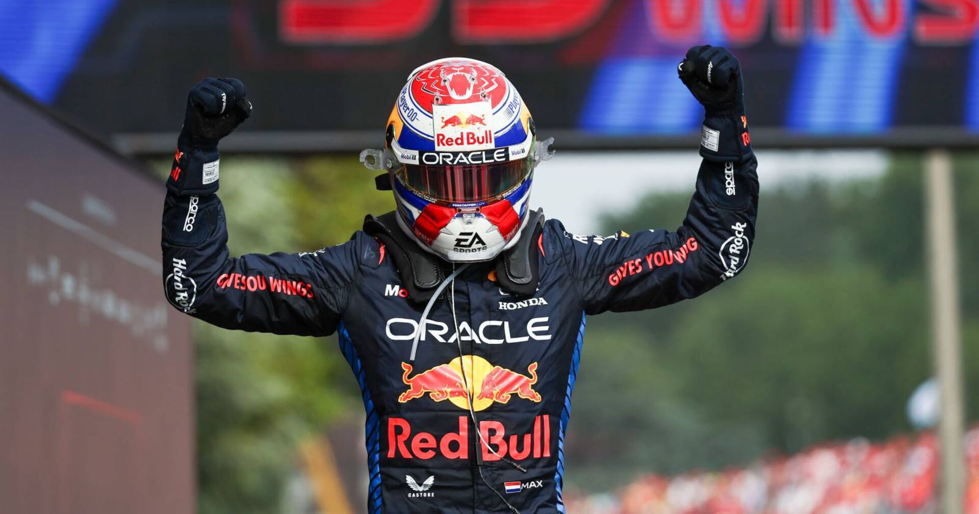 Max Verstappen, der im Zielbereich eines Grand Prix eine Bodybuilder-Geste vollführt und dabei seinen Rennhelm auf dem Kopf trägt