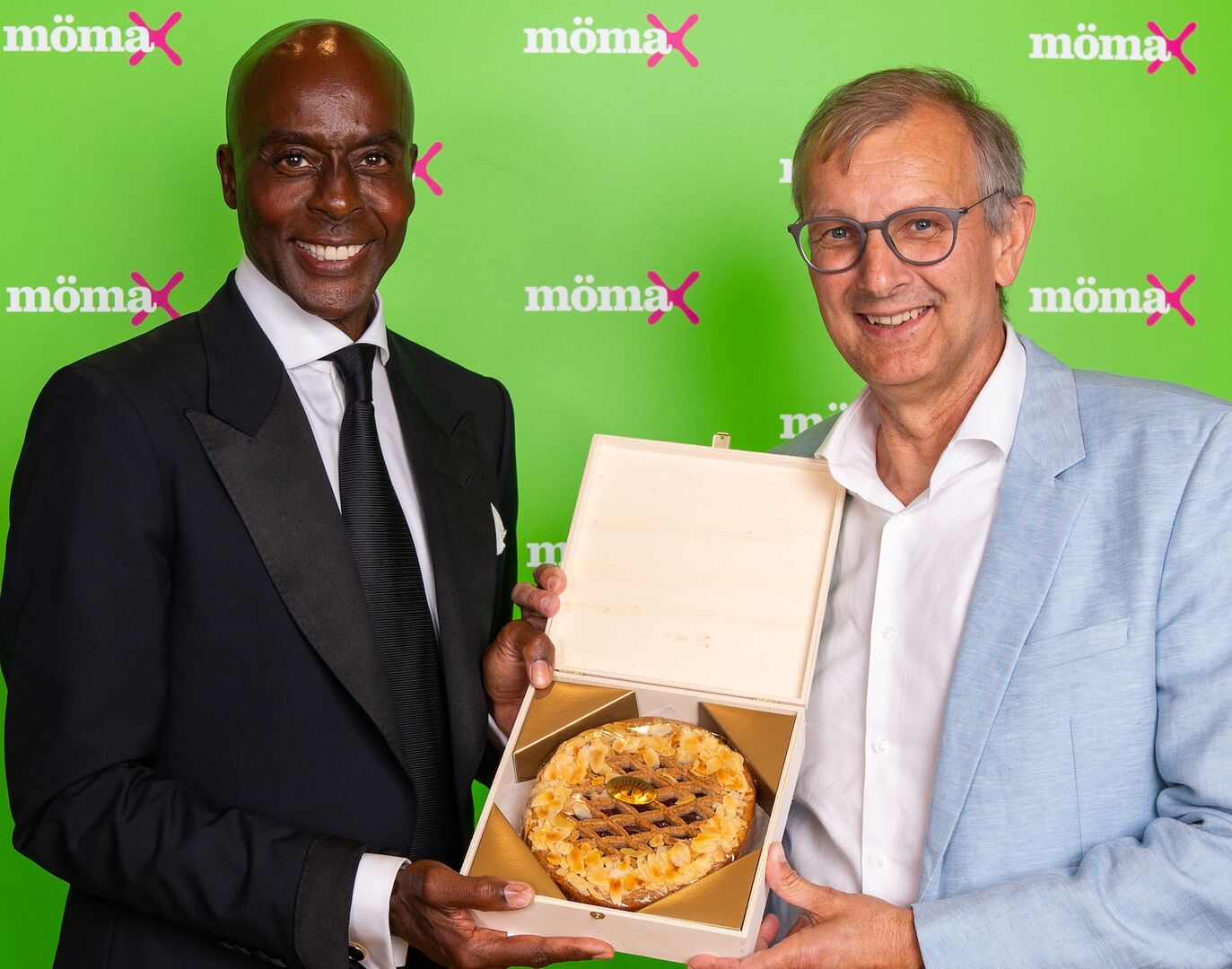 Bruce Darnell und Thomas Saliger vor einer &quot;mömax&quot;-Wand mit einer Linzer Torte in der Hand