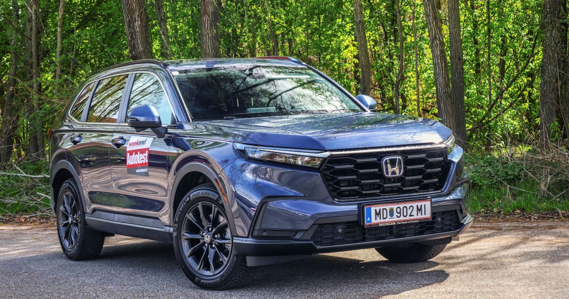 Ein blauer Honda CR-V strahlt in der Sonne. Im Hintergrund sind Laubbäume zu sehen, die ebenfalls in der Sonne leuchten.