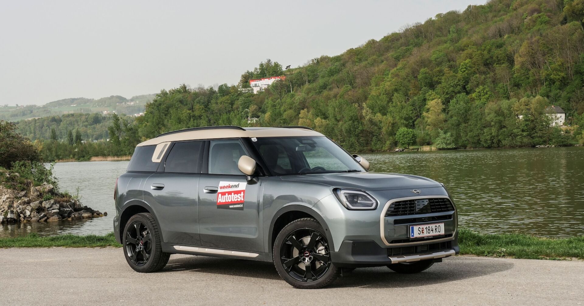 Ein silberner MINI Countryman - mit goldenen Dach - steht vor einem Fluss. Es sind keine Menschen zu sehen. Am anderen Ufer befindet sich ein dichter Laubwald, zu dem sind zwei Häuser zu sehen.