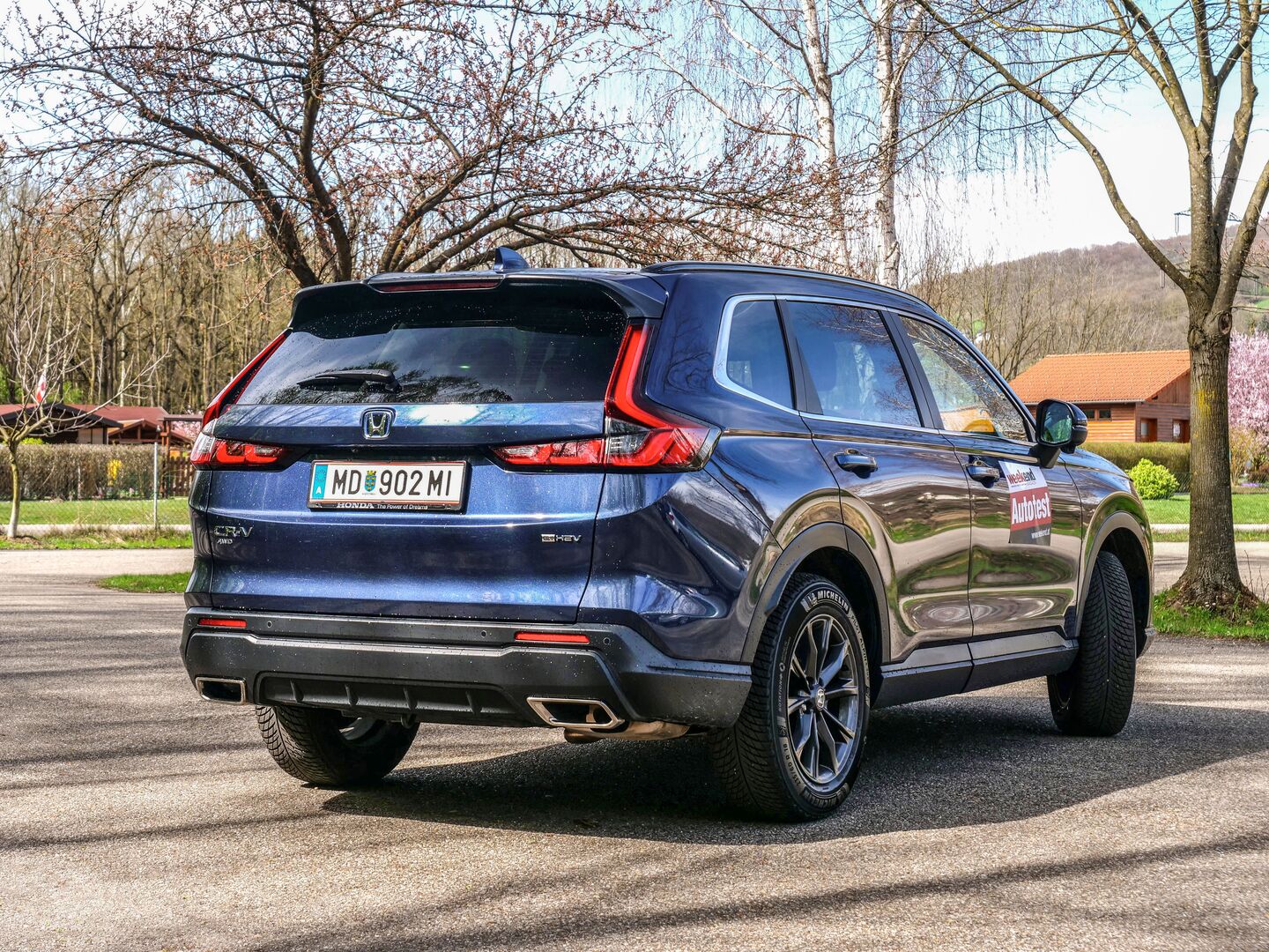 Ein blauer Honda CR-V glänzt in der Sonne. Zu sehen ist das Auto von hinten, es steht auf einem Parkplatz, im Hintergrund eine Wiese und Bäume, die noch nicht ausgetrieben sind.
