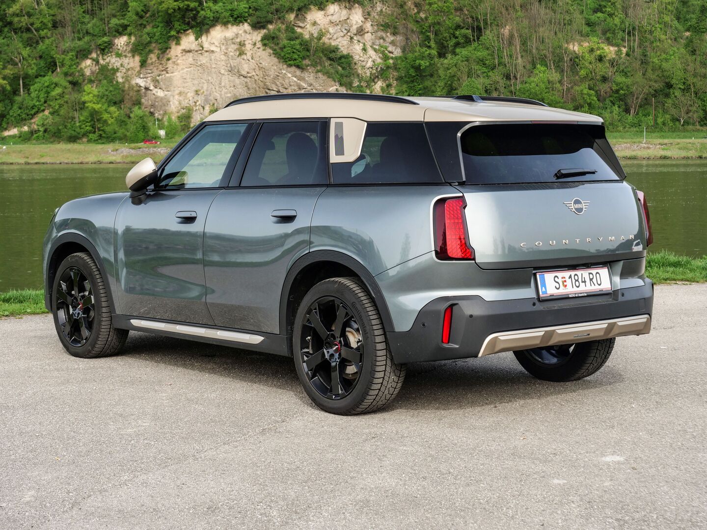 Der MINI Countryman macht von hinten eine gute Figur: Die roten Rücklichter, der silberne Lack und das goldene Dach harmonieren. Der MINI steht am Ufer eines Flusses, auf einen asphaltierten Parkplatz.
