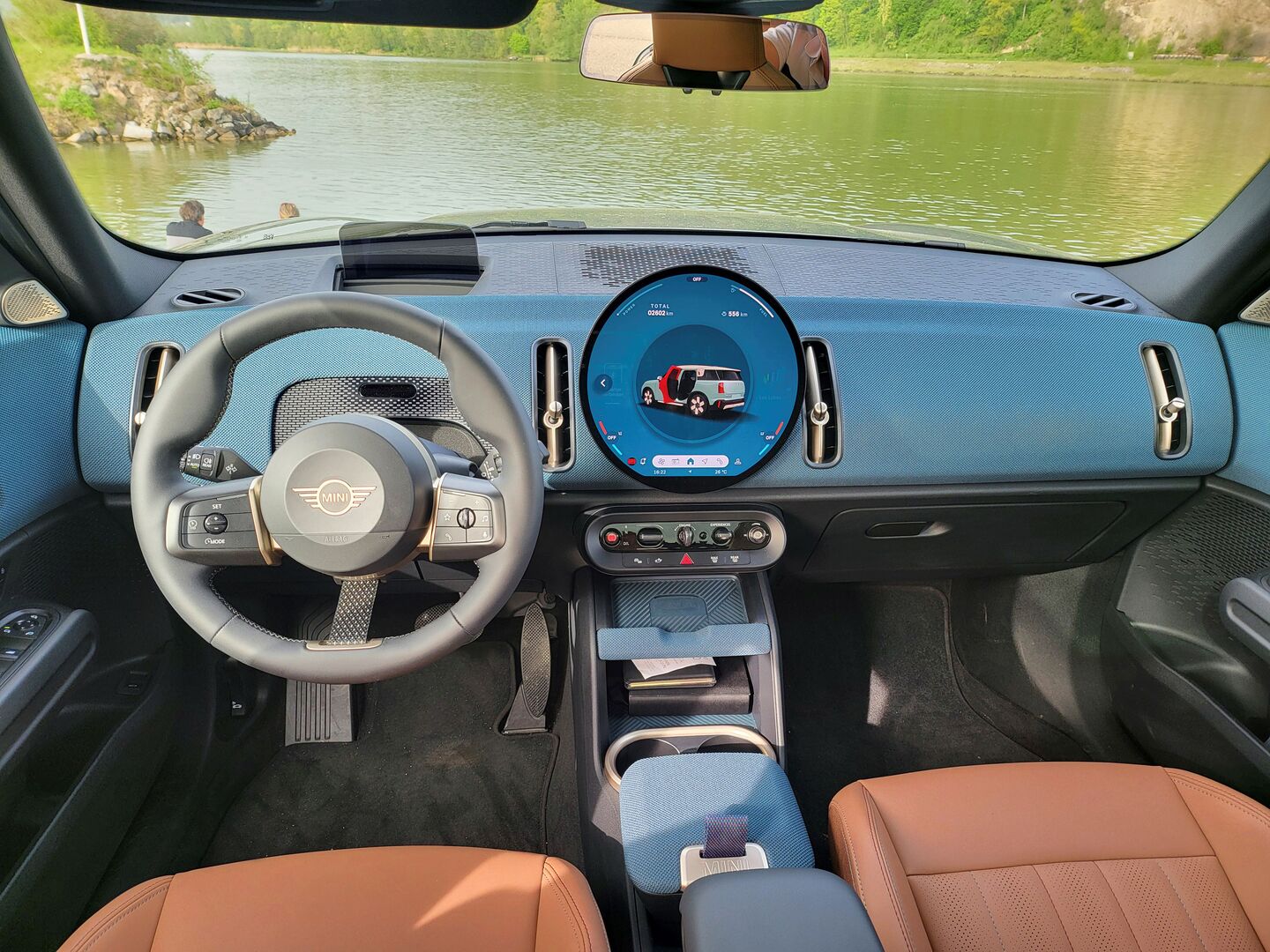 Das Innere des MINI Countryman: Braune Sitze,  das Armaturenbrett leuchtet in blaugrünem Strickmaterial. Der Screen ist rund. Knöpfe gibt es praktisch keine mehr, nur einen Drehschalter zum Starten, einen für die Fahrmodi und einen Drehknopf für die Lautstärke gibt es, dazu noch der Button für das Heizen.