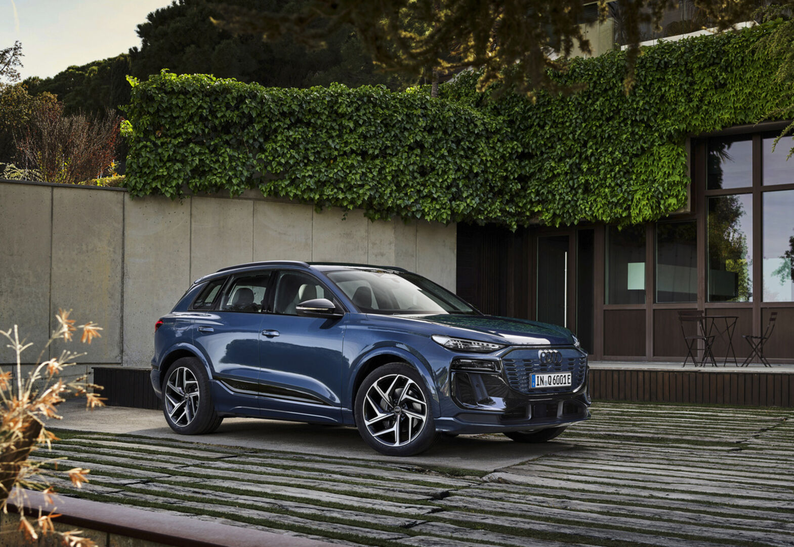 Ein blau glänzender Audi Q6 e-tron steht auf einer Terrasse. Im Hintergrund sind Efeuranken, ein Tisch und Sesseln zu sehen.