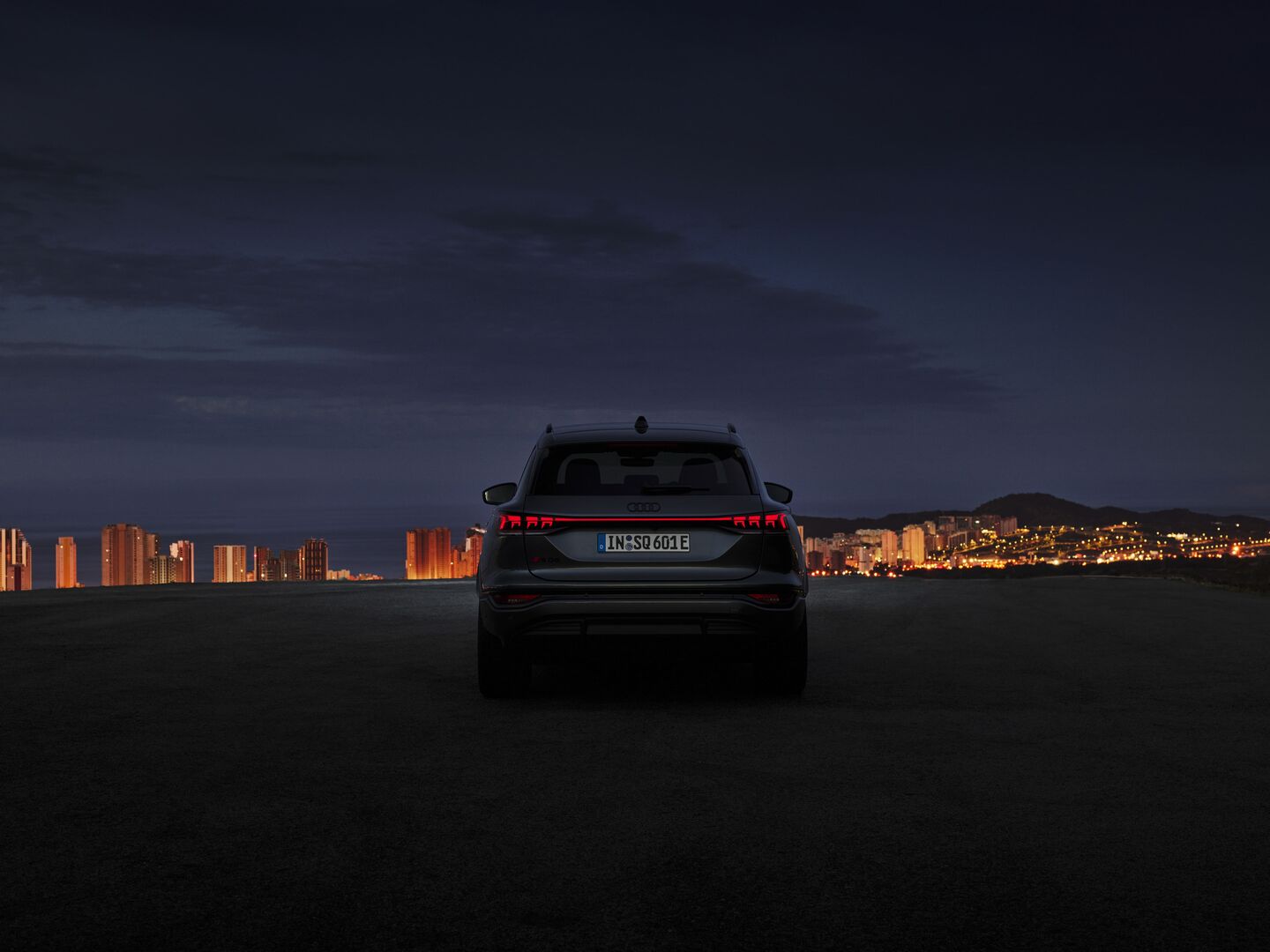 Es ist Nacht. Die Rücklichter des Audi Q6 e-tron leuchten rot. Im Hintergrund eine Großstadt am Meer. Die Häuser sind beleuchtet.