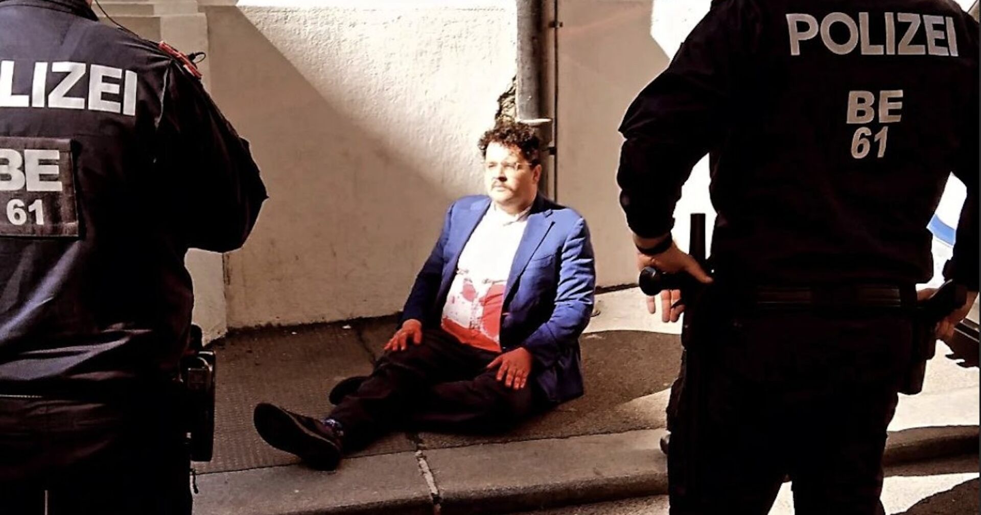 Der Aktivist, der die Ministerin attackiert hat, sitzt mit Kunstblut befleckt am Boden. Die Polizei bewacht ihn.