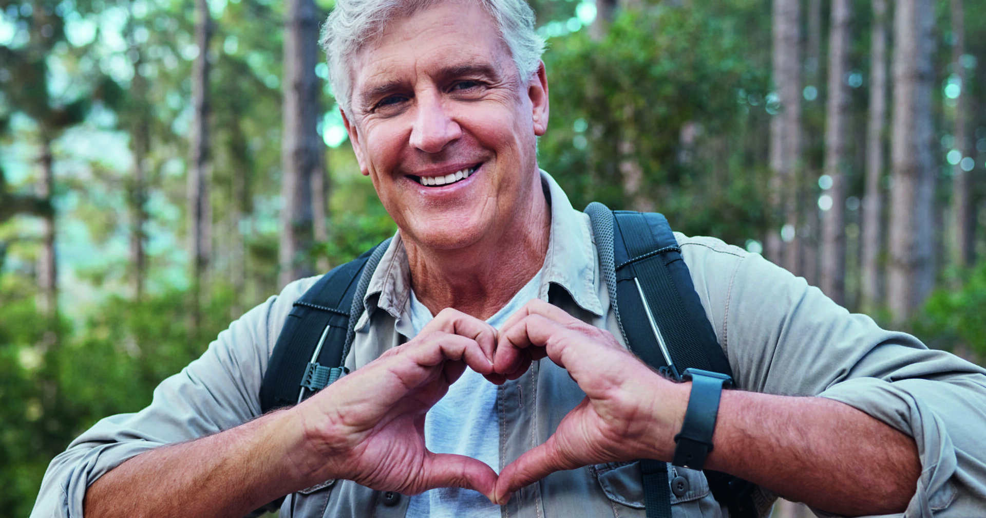 Ein Mann mit weißen Haaren steht in einem Nadelwald. Er trägt eine hellgrüne Jacke und einen Rucksack. Der Mann lacht und hat seine beiden Hände vor sich zu einem Herz geformt.