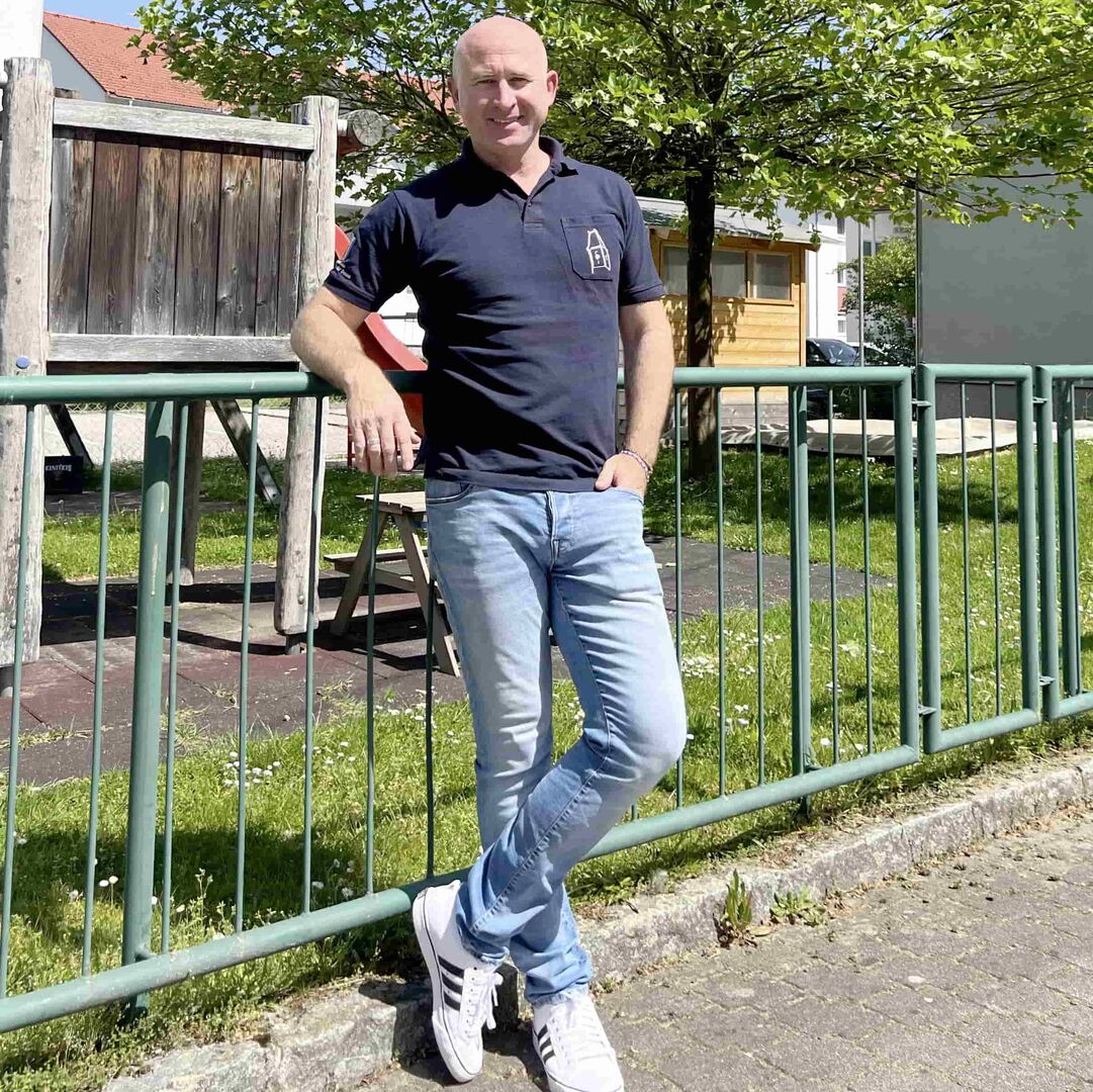 Bürgermeister von Feldkirchen, David Allerstorfer, trägt weiße Turnschuhe, ein hellblaue Jean und ein dunkelblaues T-Shirt. Der Mann - mit Glatze - steht vor dem Zaun des Kindergartes und lacht.