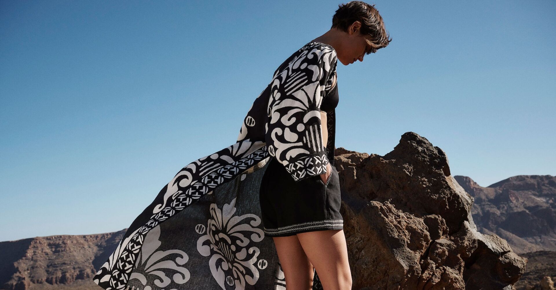 Model mit flatterndem Mantel und Shorts vor Felsenkulisse