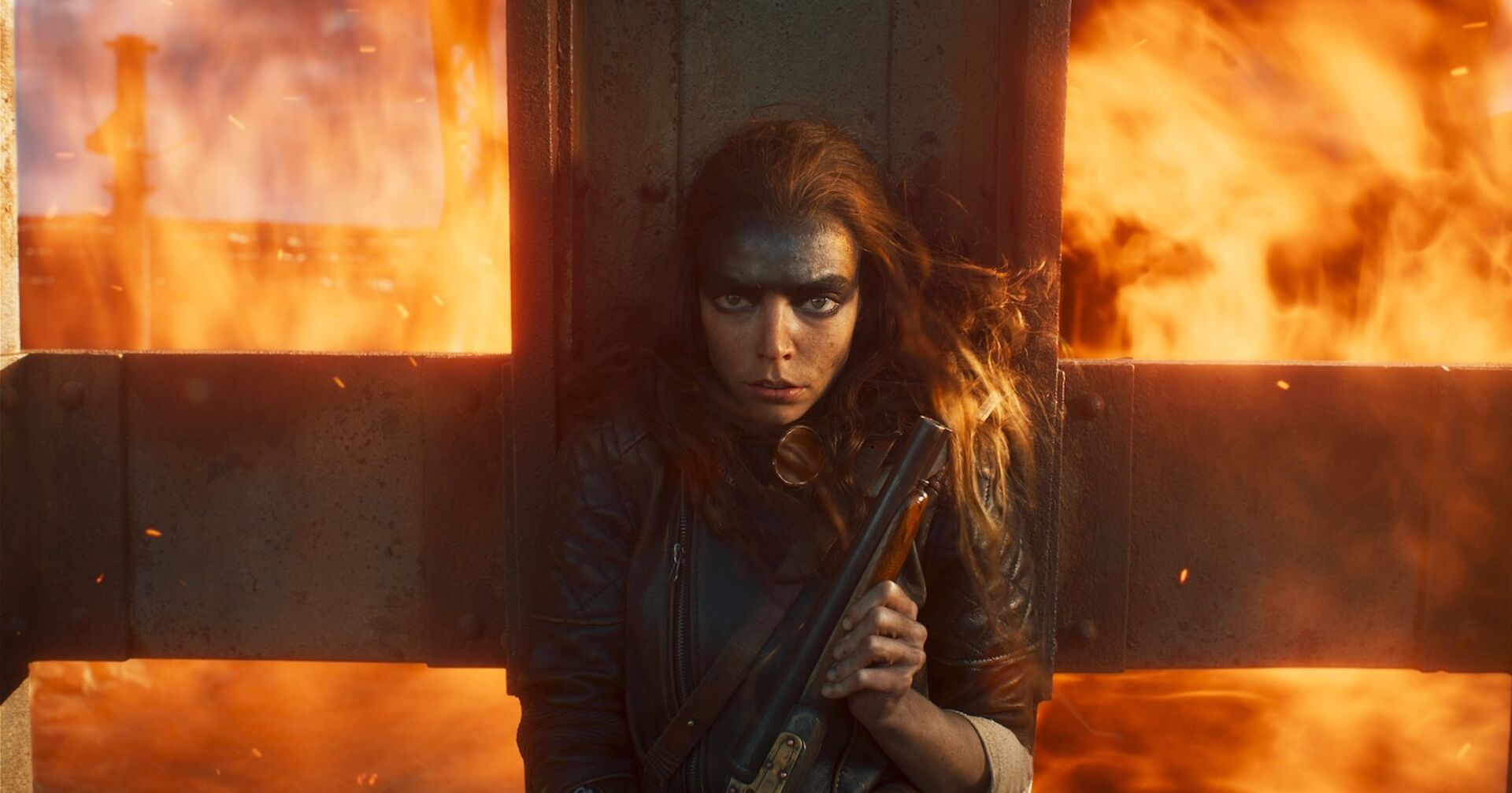 Eine Schauspielerin vom Film &quot;Furiosa: A Mad Max Saga&quot; mit viel schwarzer Schimnke. Hinter ihr sind Flammen. Sie trägt eine Waffe.