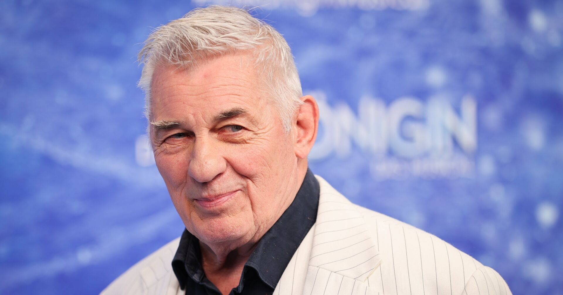 Heinz Hoenig vor einer hellblauen Wand mit einem weißen Sakko und einem schwarzen Hemd.