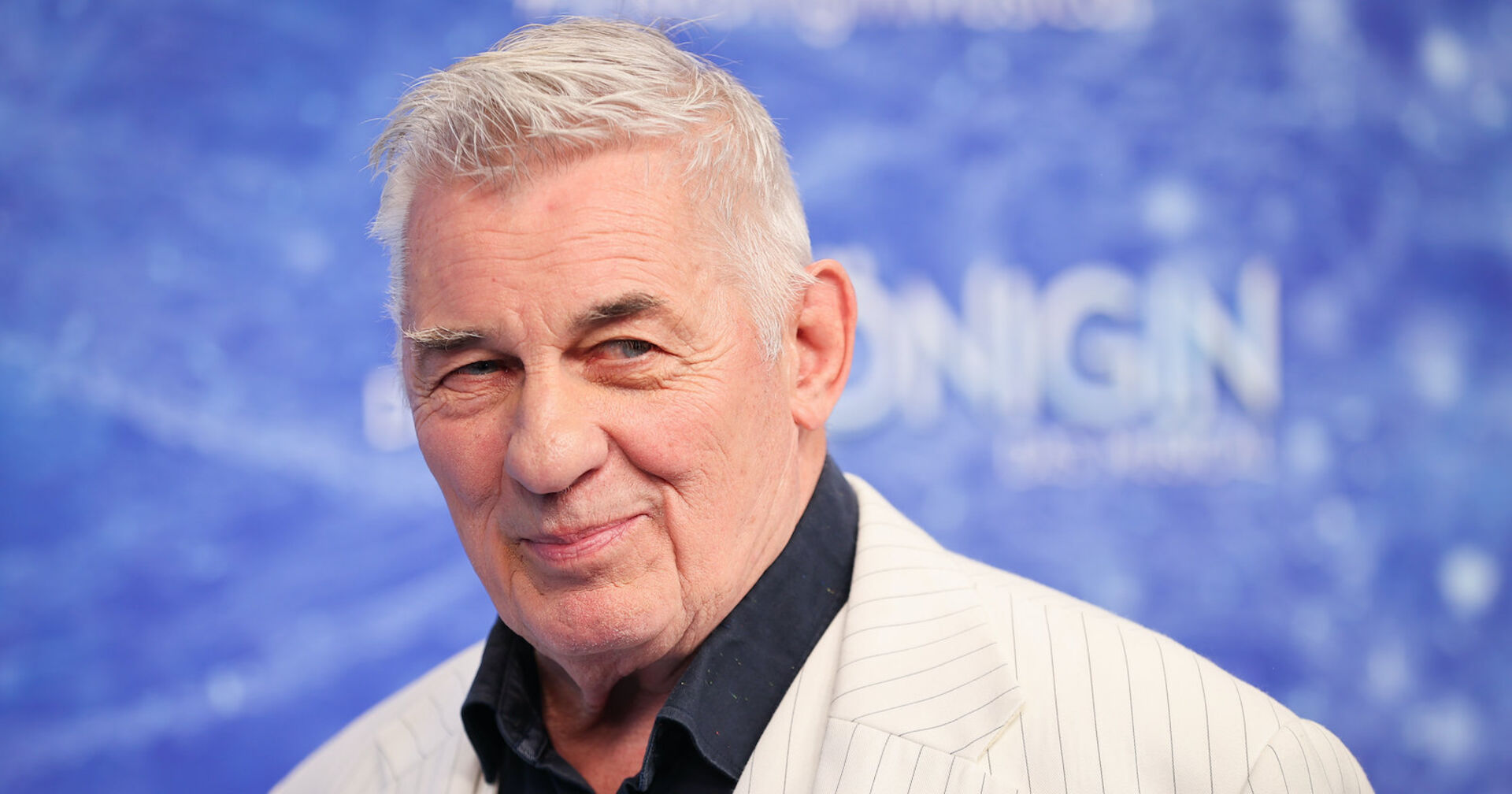 Heinz Hoenig mit eiem weißen Sakko und einem schwarzen Hemd vor einer blauen Fotowand.