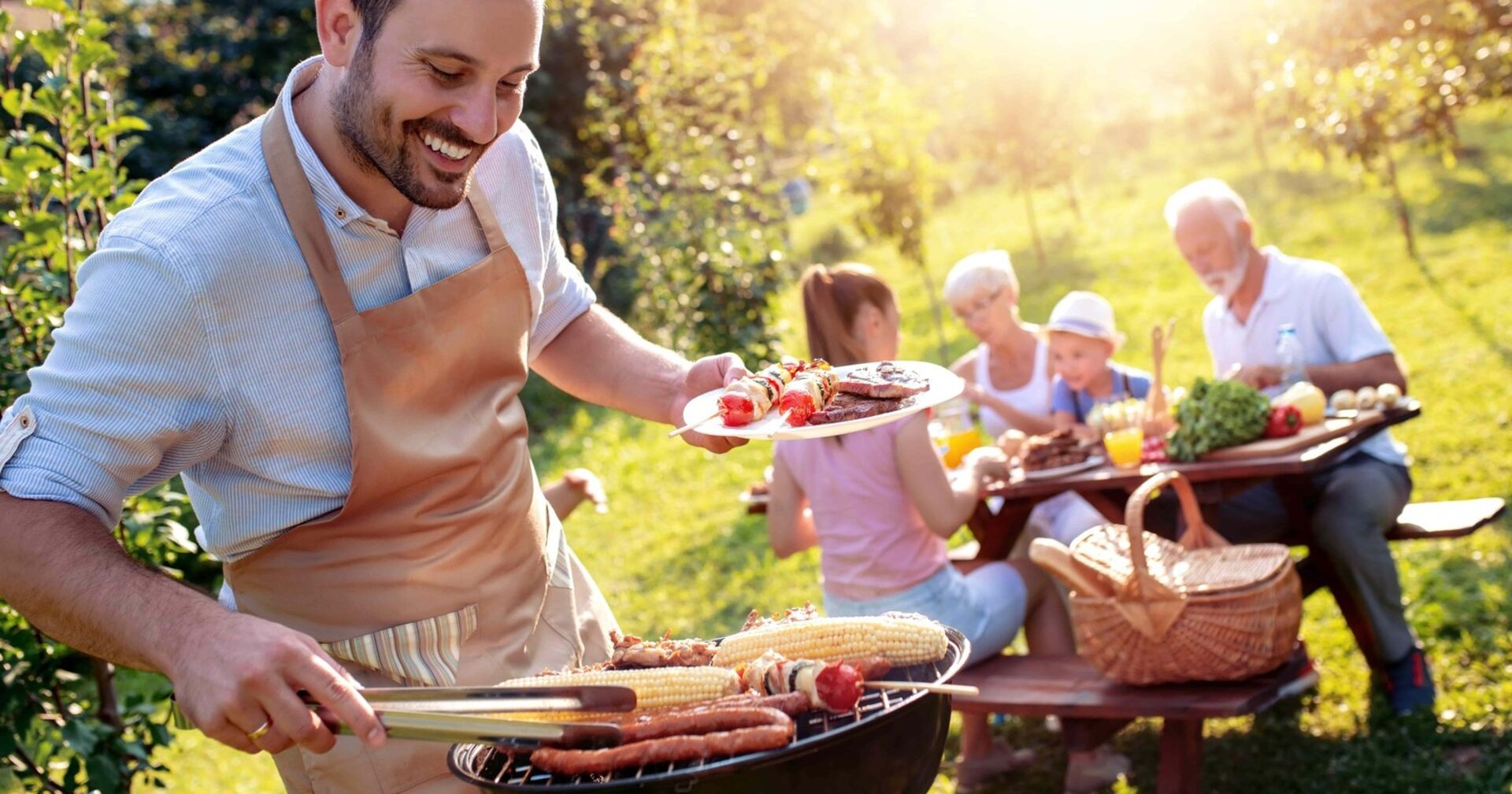 Ein Mann mit schwarzem Vollbart - bekleidet mit einer brauner Schürze grill. In der linken Hand hält er einen Teller mit gegrillten Fleisch und Gemüse, mit der rechten Hand wendet er mithilfe eine Zange einen Maiskolben, der am Grill liegt. Die Sonne scheint, im Hintergrund essen zwei Kinder, eine Frau und ein ältere Herr die gegrillten Speisen.