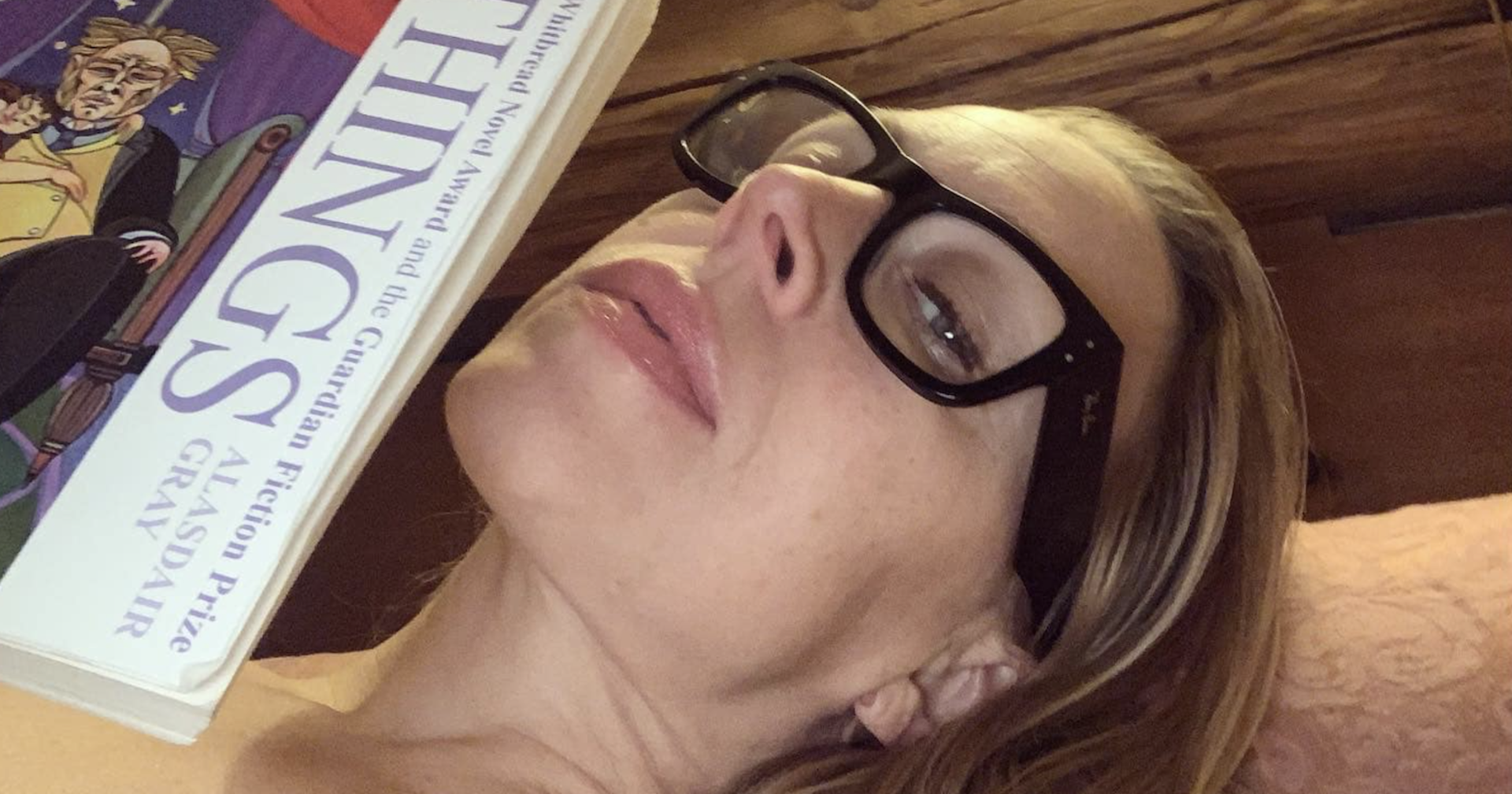 Nina Proll mit Brille räkelt sich auf einem Bett, in der Hand hat sie ein Buch