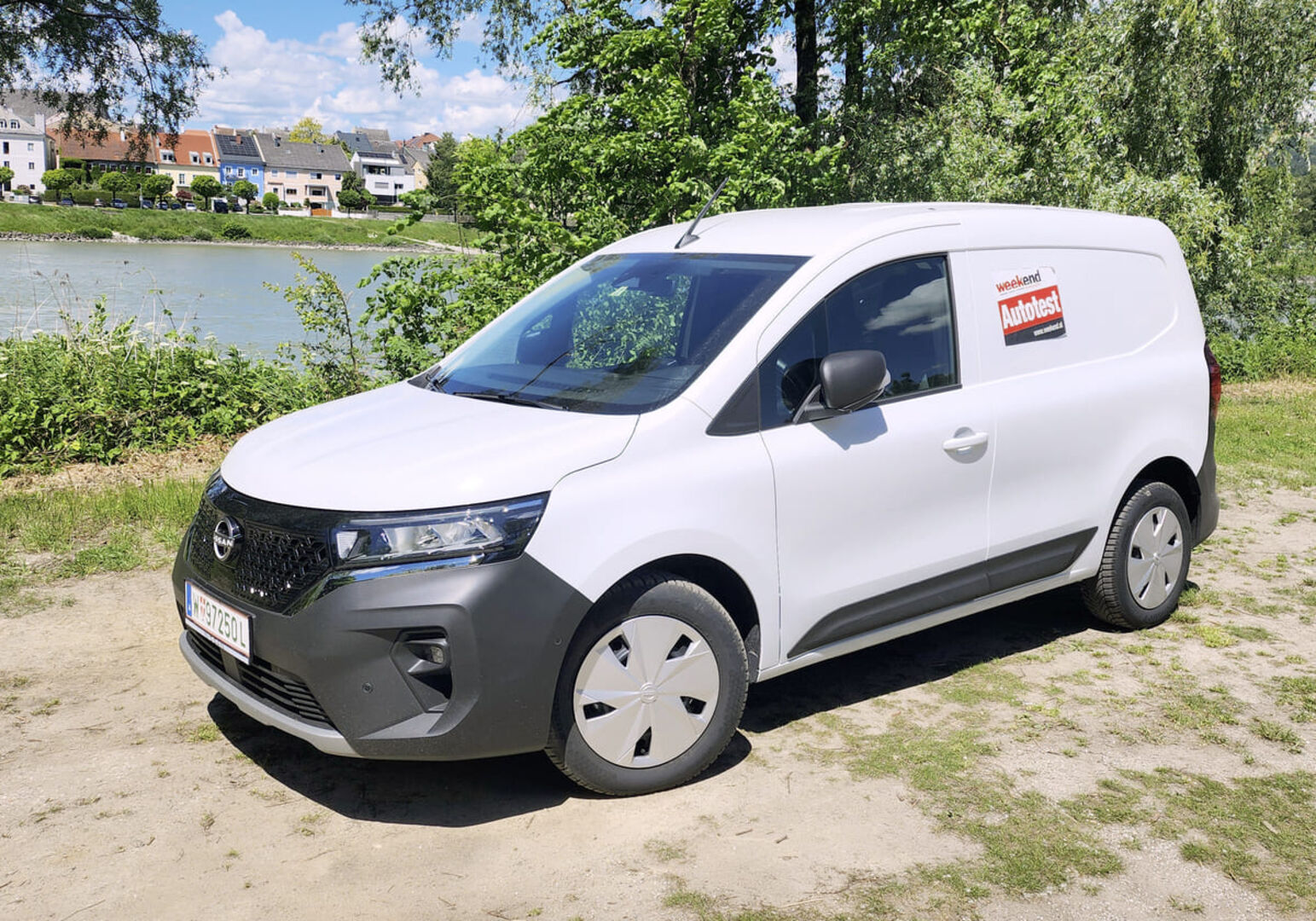 Ein weißer Nissan Townstar glänzt in der Sonne. Das Auto steht am Ufer eines Flusses, auf einem sandigen Parkplatz. Am anderes Ufer sind mehrere Häuser zu sehen.