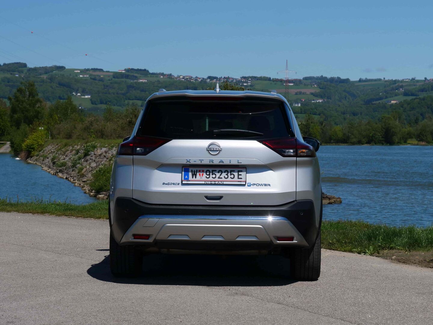 Das silberne Heck des Nissan X-Trail glänzt in der Sonne. Das Auto steht auf einem asphaltierten Parkplatz. Im Hintergrund ist ein Fluss und grüne Hügel zu sehen. In der Ferne sind auch einige Häuser zu erkennen.