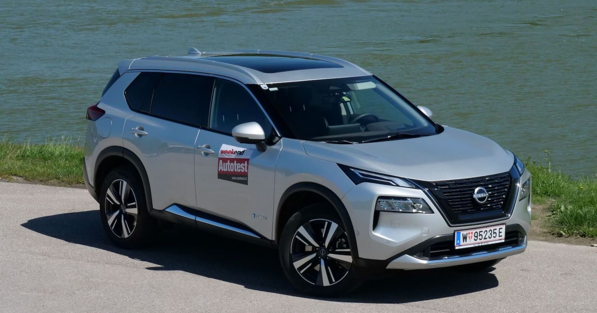 Ein silberner Nissan X-Trail glänzt in der Sonne. Er steht am Ufer eines Flusses auf einem asphaltierten Parkplatz.