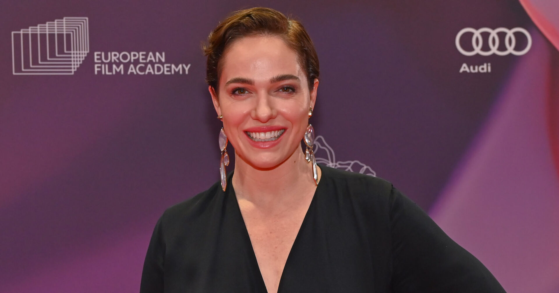 Verena Altenberger mit einem schwarzen Kleid vor einer violetten Fotowand.