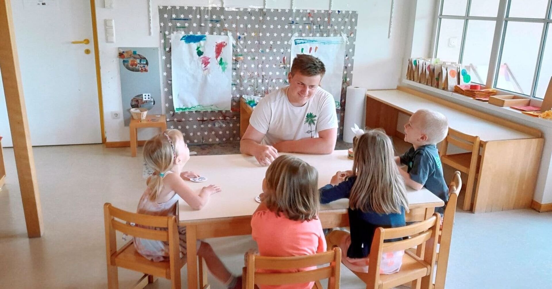 Zivildiener Julian Schober trägt ein weißes T-Shirt. Er sitzt mit vier Kindern an einem braunen Holztisch und Holzsesseln. Der Raum im Kindergarten von Feldkichen an der Donau ist hell und mit einigen von Kindern bemalten Plakaten geschmückt.