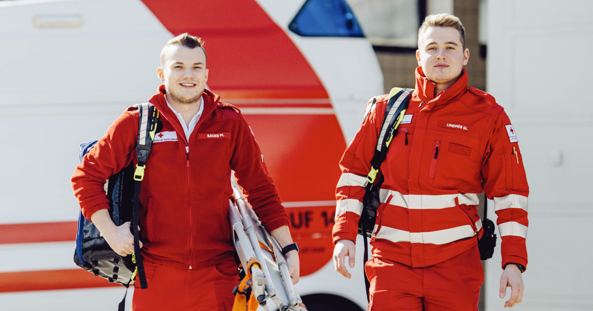 Zwei Junge Männer beim Roten Kreuz: Beide sind mit der roten Uniform des Roten Kreuzes bekleidet. Der Linke trägt einen blauen Rucksack und eine zusammengeklappte Trage. Der rechte nur einen Rucksack, der kaum zu sehen ist. Im Hintergrund wartet das Rettungsfahrzeug.