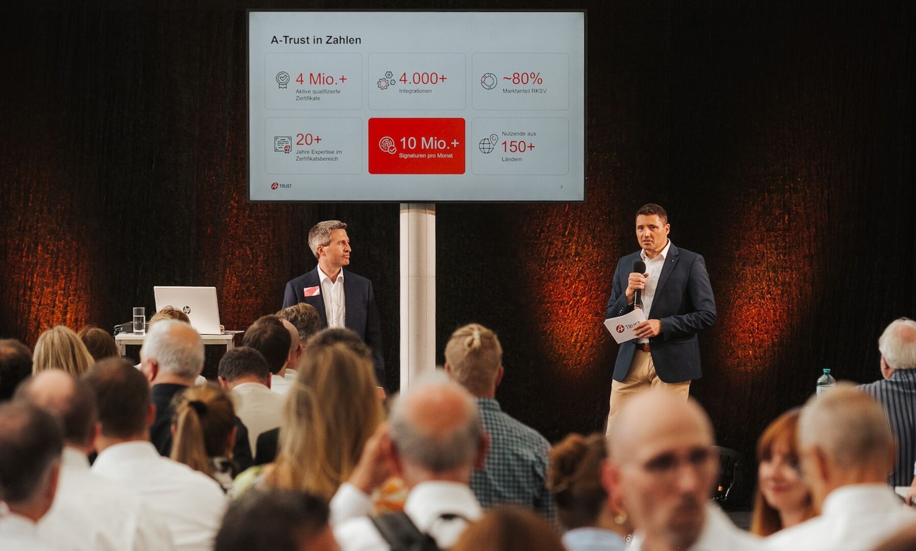 Zwei Männer mit Whiteboard am Podium, davor Publikum