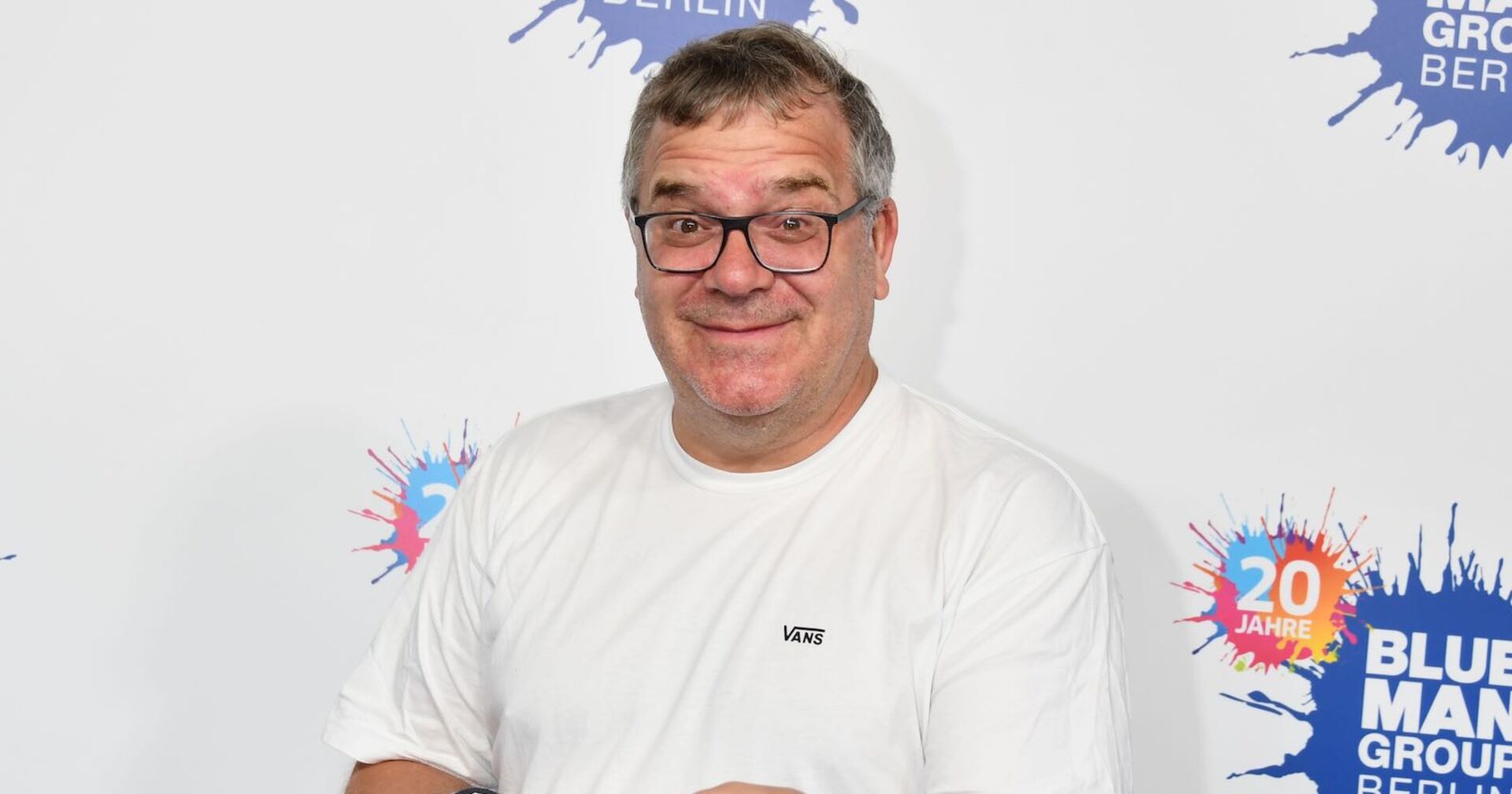 Moderator Elton vor einer Fotowand mit einem weißen T-Shirt.