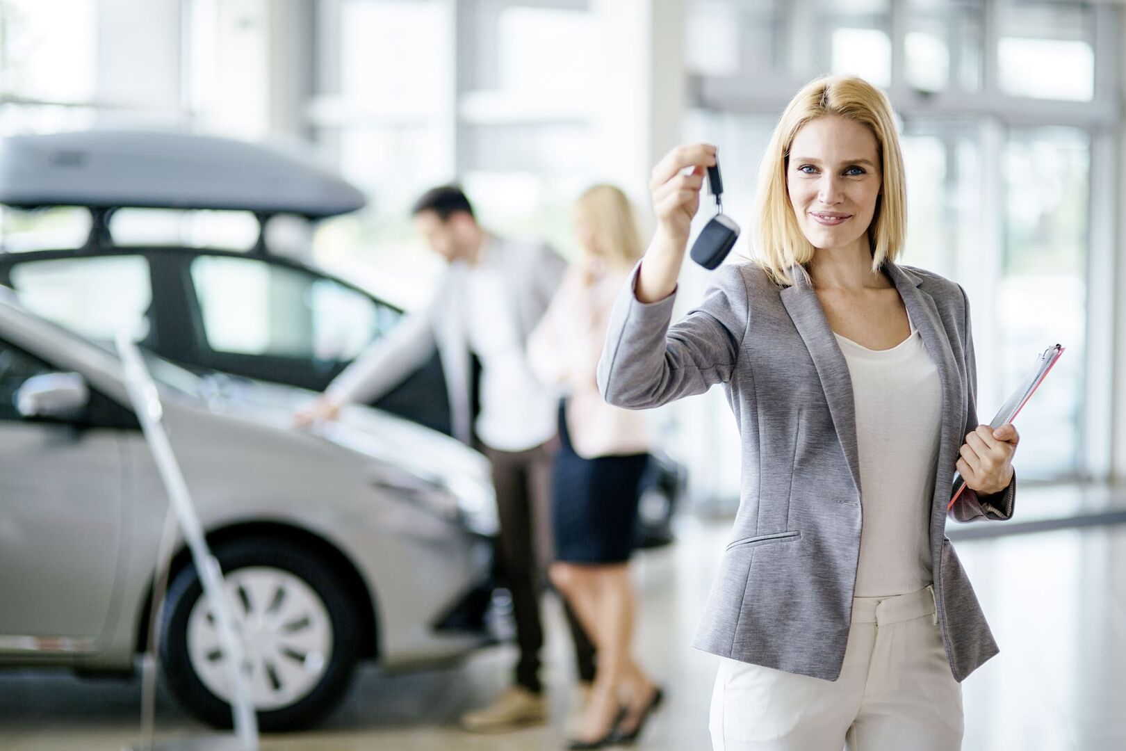 Eine blonde Frau hält in der einen Hand einen Autoschlüssel, in der anderen eine rote Mappe. Die Dame trägt eine weiße Hose, eine weißes T-Shirt und eine grau-blaues Sakko. Im Hintergrund sehen sich eine Mann und eine Frau ein neues Auto an.