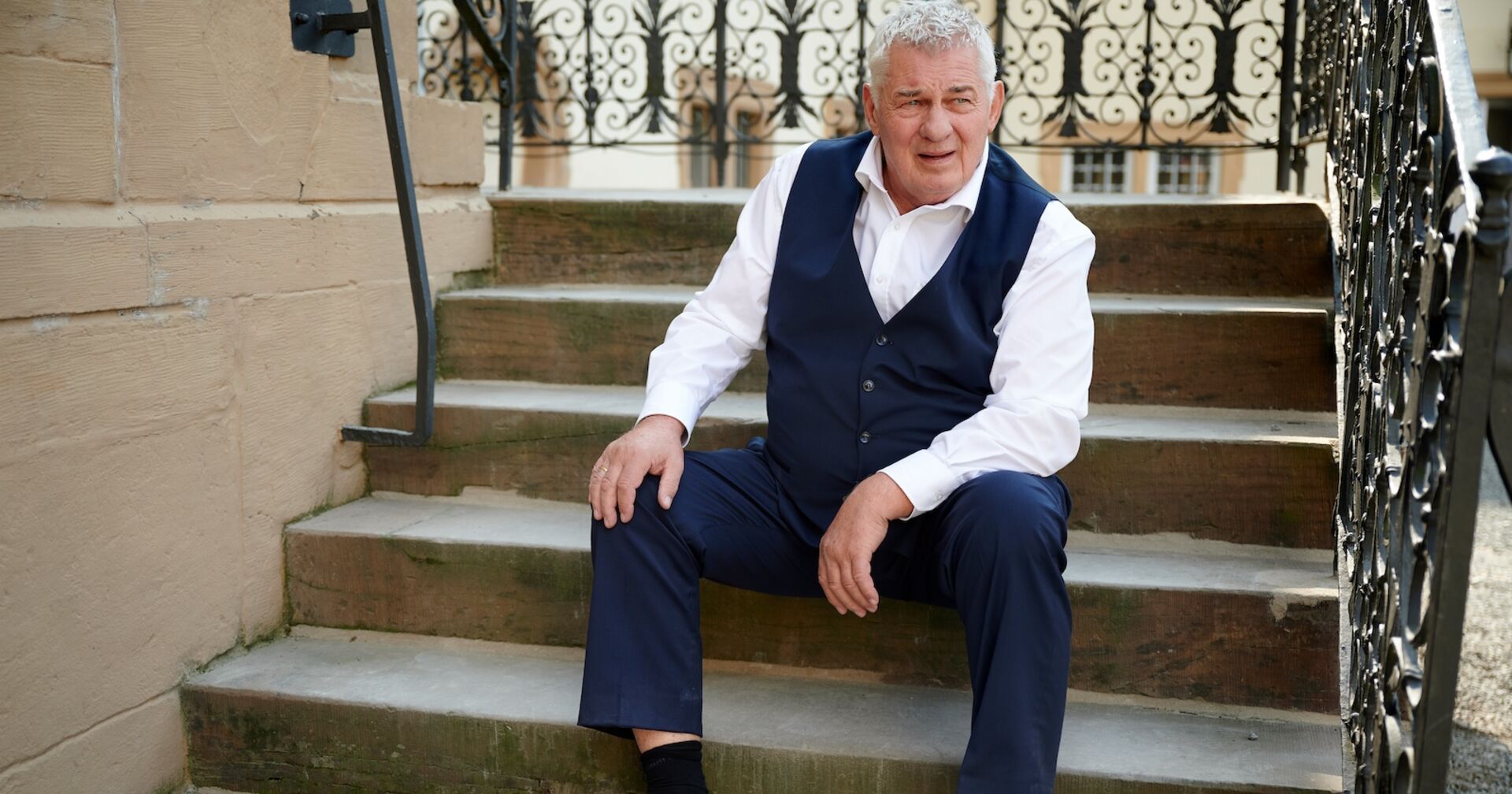 Heinz Hoenig in einem dunkelblauen Sakko und einem weißen Hemd auf Treppen sitzend.