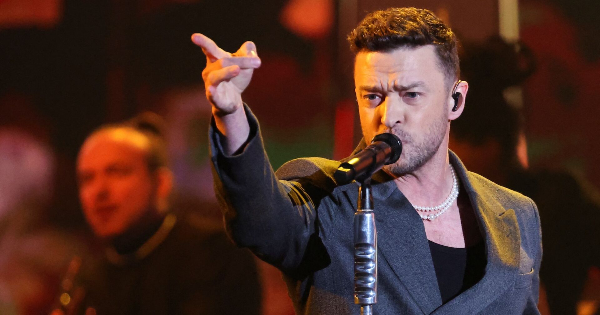 Justin Timberlake mit einem grauen Sakko vor einem Mikrofon. Er zeigt ins Publikum.