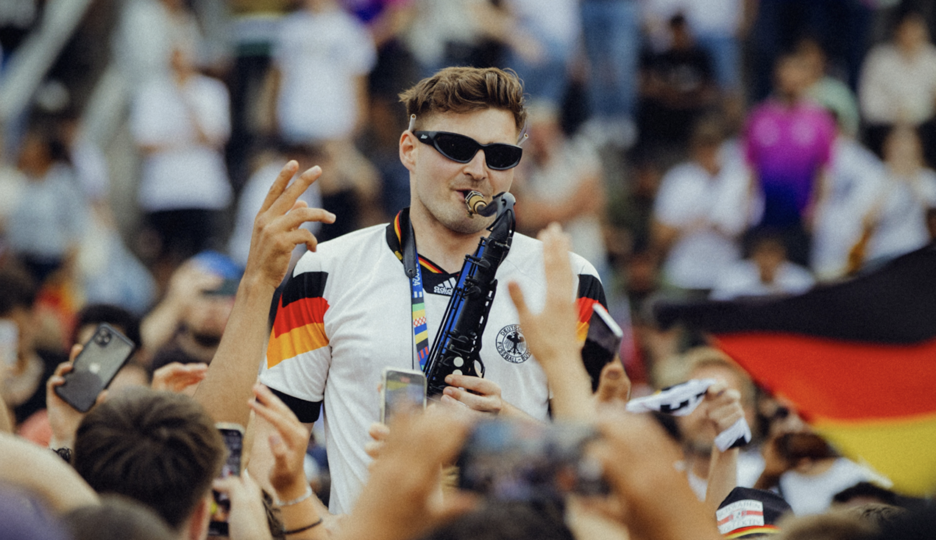 Ein Mann trägt eine Sonnenbrille und spielt ein Saxophon, während ihm alle zujubeln