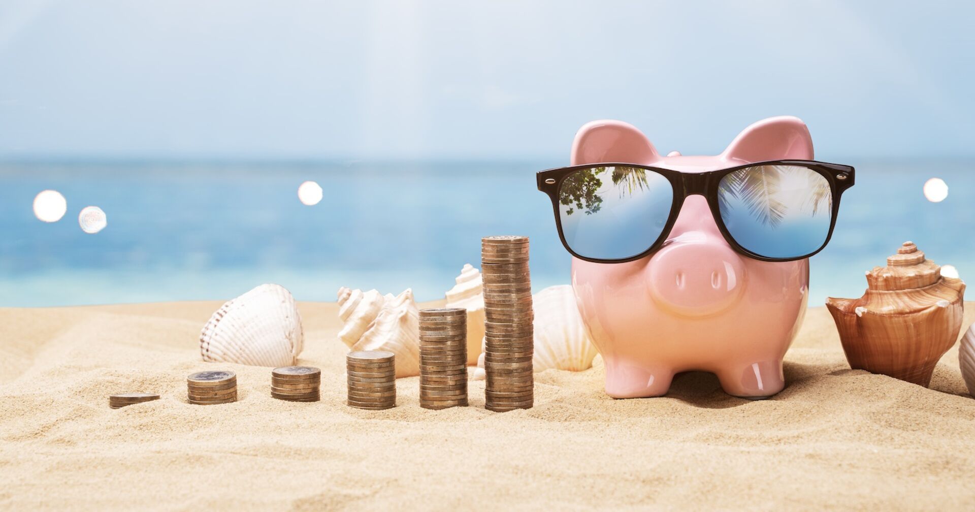 Ein Sparschwein am Strand, daneben Münzstapel