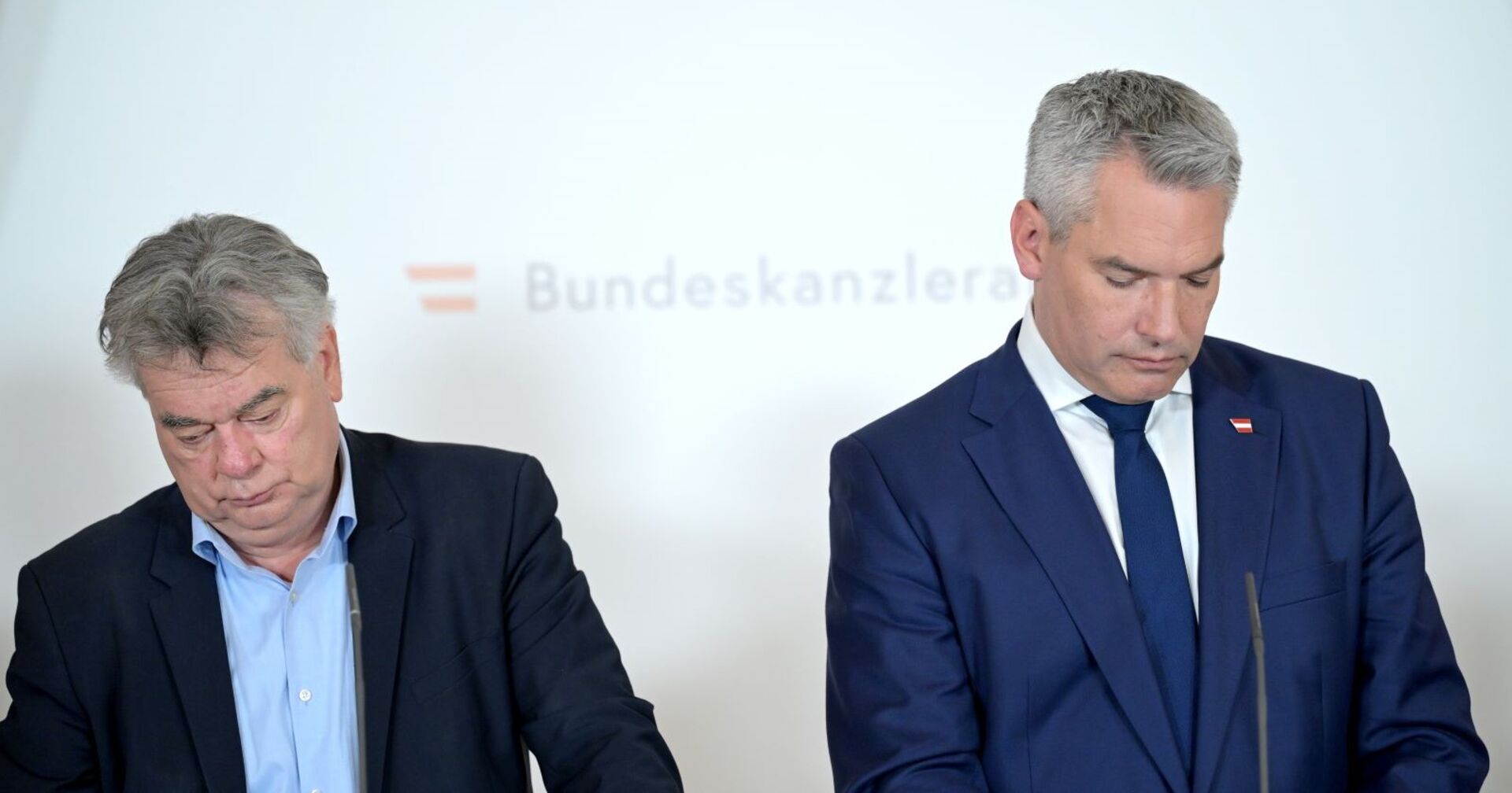 Werner Kogler und Karl Nehammer bei einer Pressekonferenz im Bundeskanzleramt. Sie stehen leicht abgewandt zueinander und schauen nach unten auf ihre Notizen.
