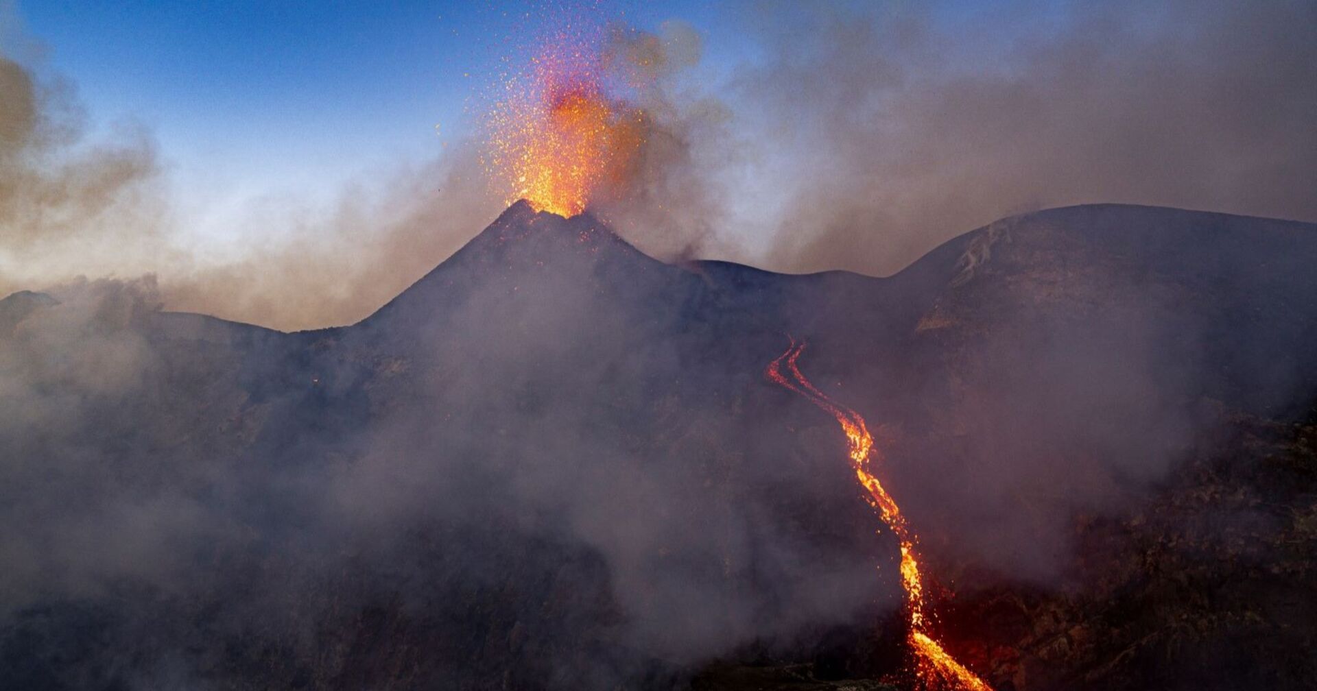 Der Ätna bei Dämmerung spuckt Lava und Rauchwolcken, an der Seite des Vulkan fließt Lava herab