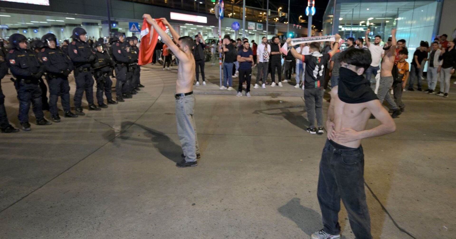 Ein vermummter Jugendlicher mit nackten Oberkörper steht vor einer Phalanx Polizisten mit Schutzschildern, hinter ihm weitere Jugendliche