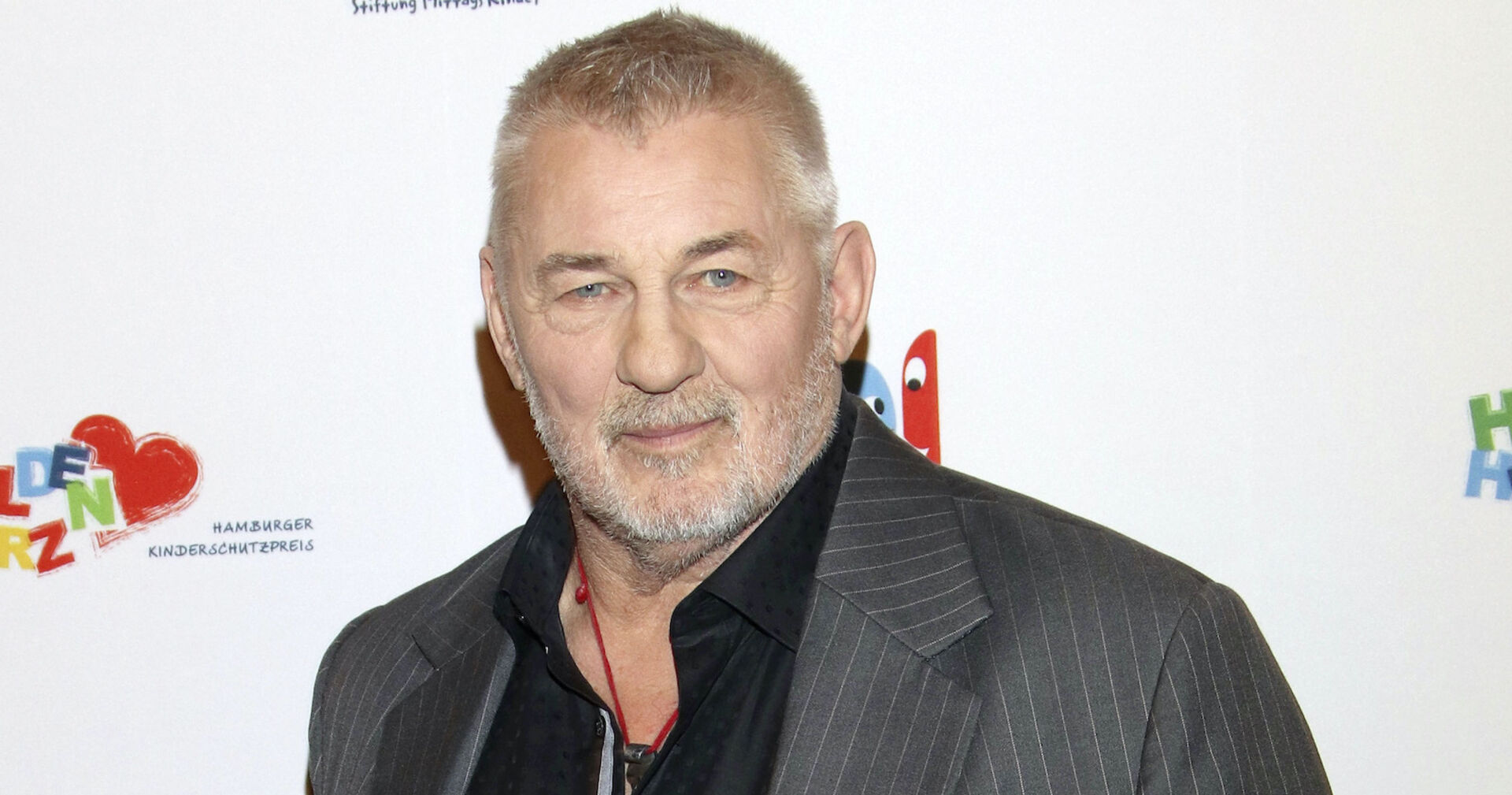 Heinz Hoenig mit einem grauen Sakko vor einer Fotowand.