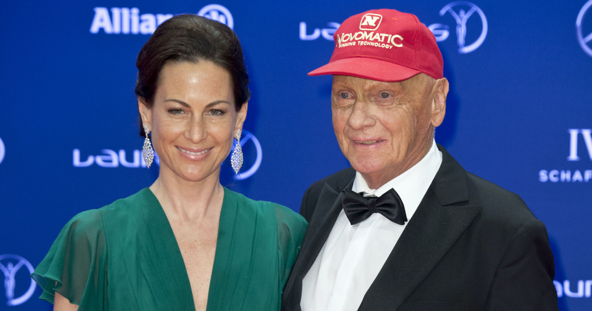 Niki und Birgit Lauda auf einem Event vor einer blauen Fotowand. Sie trägt ein grünes Kleid und Niki Lauda einen Smoking und eine rote Kappe.