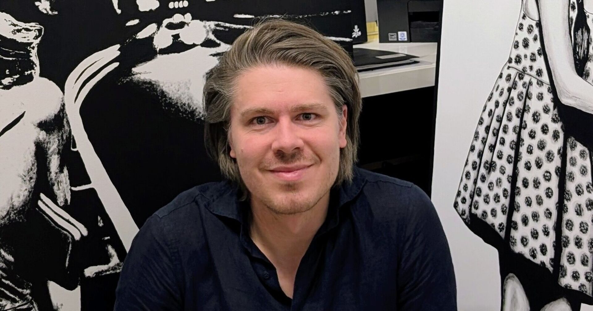 Der Künstler Jürgen Vogel lacht in die Kamera. Er trägt ein blaues Hemd, hat lange braune Haare und einen Drei-Tages-Bart. Im Hintergrund sind Bilder in Schwarz-Weiß zu erkennen.