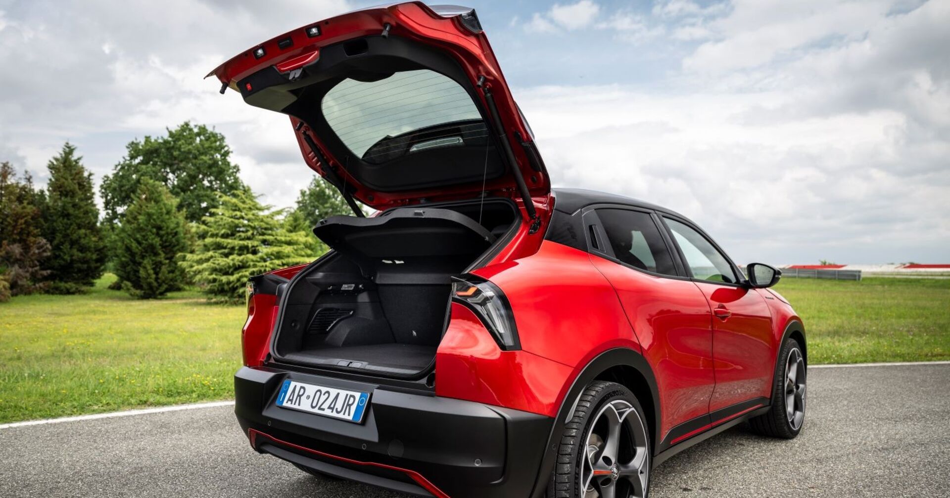 Ein roter Alfa Romeo Junior von hinten. Der Kofferraum ist geöffnet und man man sieht die schwarte Ladefläche. Das Auto steht auf einer asphaltierten Straße, im Hintergrund sind kleine Bäume und eine Wiese.