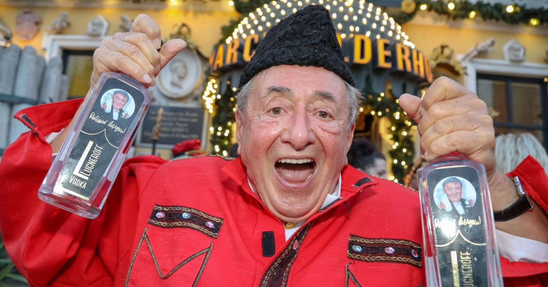 Richard Lugner in rotem Hemd und schwarzem Hut mit zwei Wodkaflaschen in seiner Hand