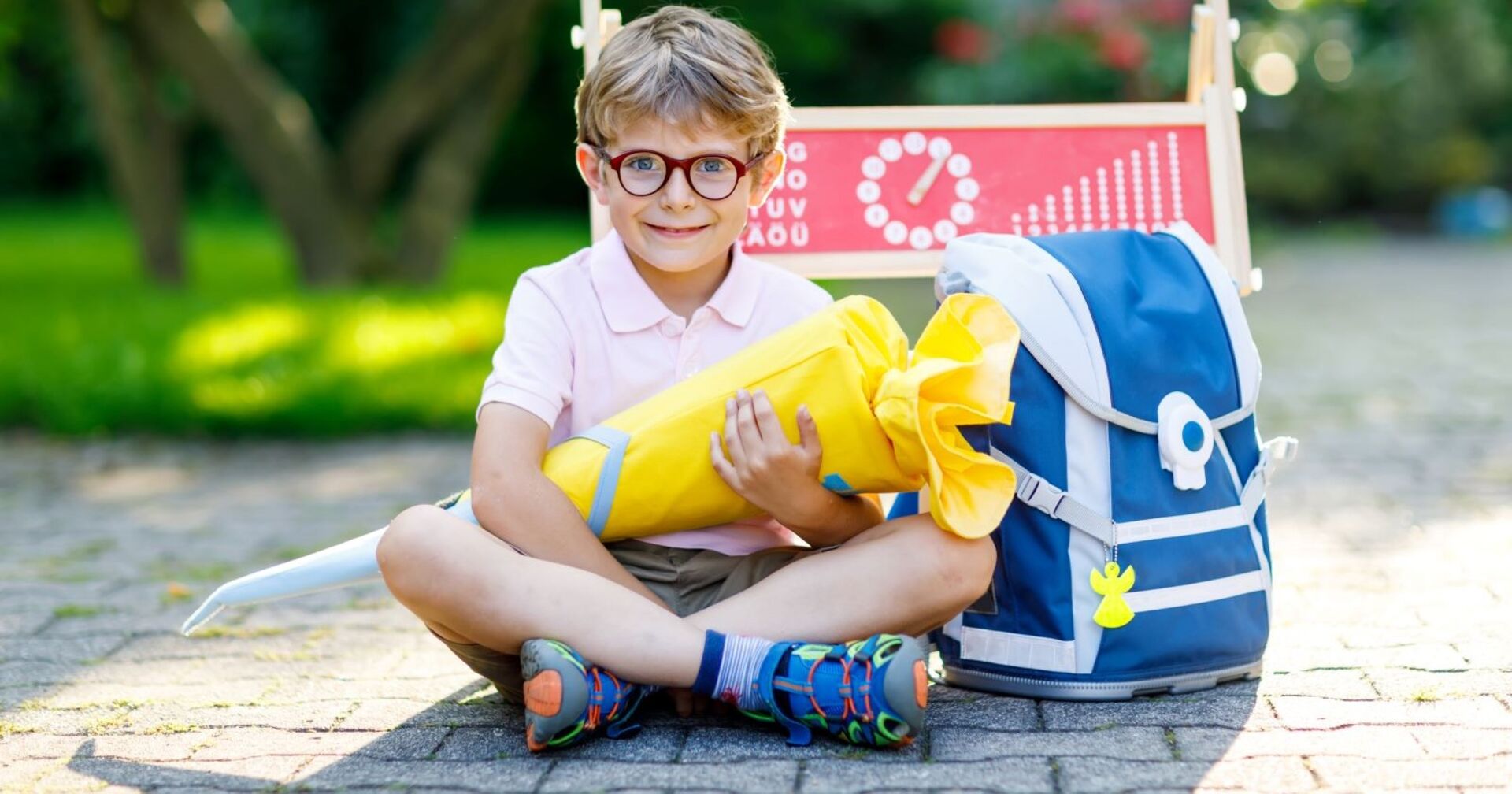 Ein junger Bub - mit kurzer Hose, kurzem Polo und bunten Turnschuhen bekleidet - lacht in die Kamera. Er trägt eine rote Brille und in der Hand hält er eine gelbe Schultüte. Rechts von ihm steht eine blaue Schultasche, hinter ihm eine Kreidetafel auf der Schule zu lesen ist.