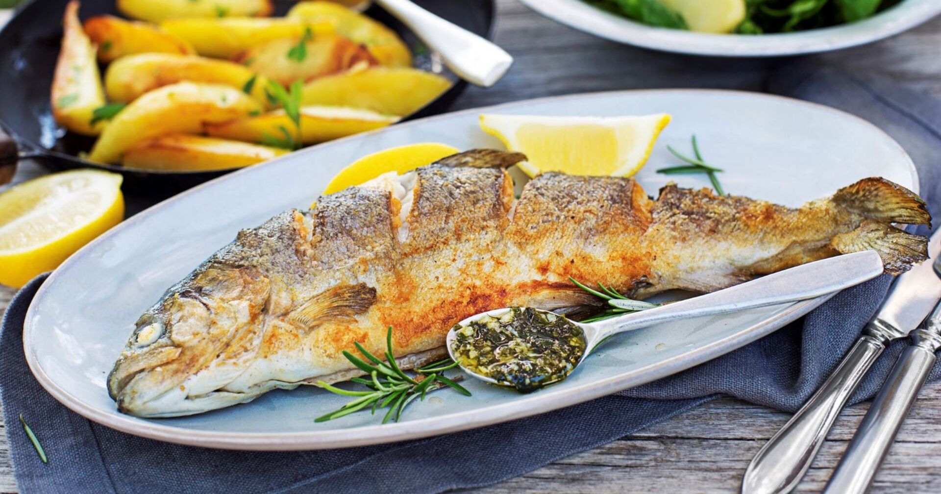 Ein kross gebratener Fisch - wahrscheinliche ein Forelle - liegt auf einem weißen Teller. Als Dekoration dienen Zitronenspalten und Rosmarinzweige. Im Hintergrund steht ein Teller mit Erdäpfeln. Zu sehen ist auch Besteck und eine Serviette.