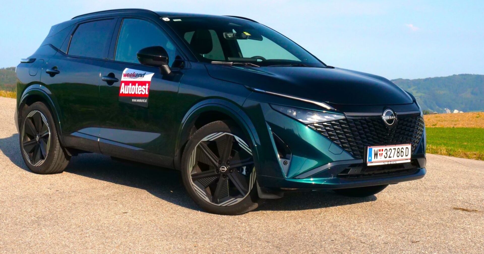 Ein dunkelgrüner Nissan Qashqai steht auf einem asphaltierten Parkplatz. Im Hintergrund sind Felder und einige Hügel zu sehen. Das Auto glänzt in der Sonne.