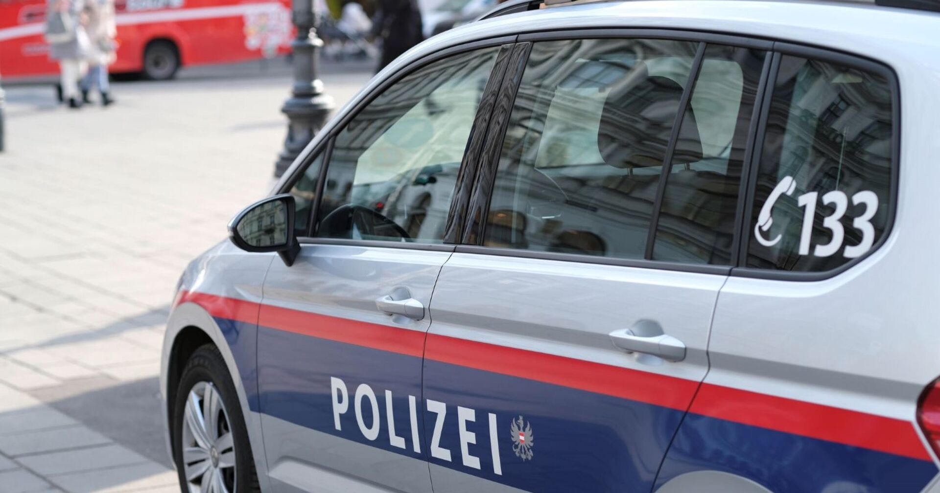 Ein unbesetztes Auto der österreichischen Polizei steht auf einem asphaltierten Platz. Auf der Seite des Autos ist die Telefonnummer 133 und die Aufschrift Polizei zu lesen. Im Hintergrund sind ein roter Bus und einige Menschen zu sehen.