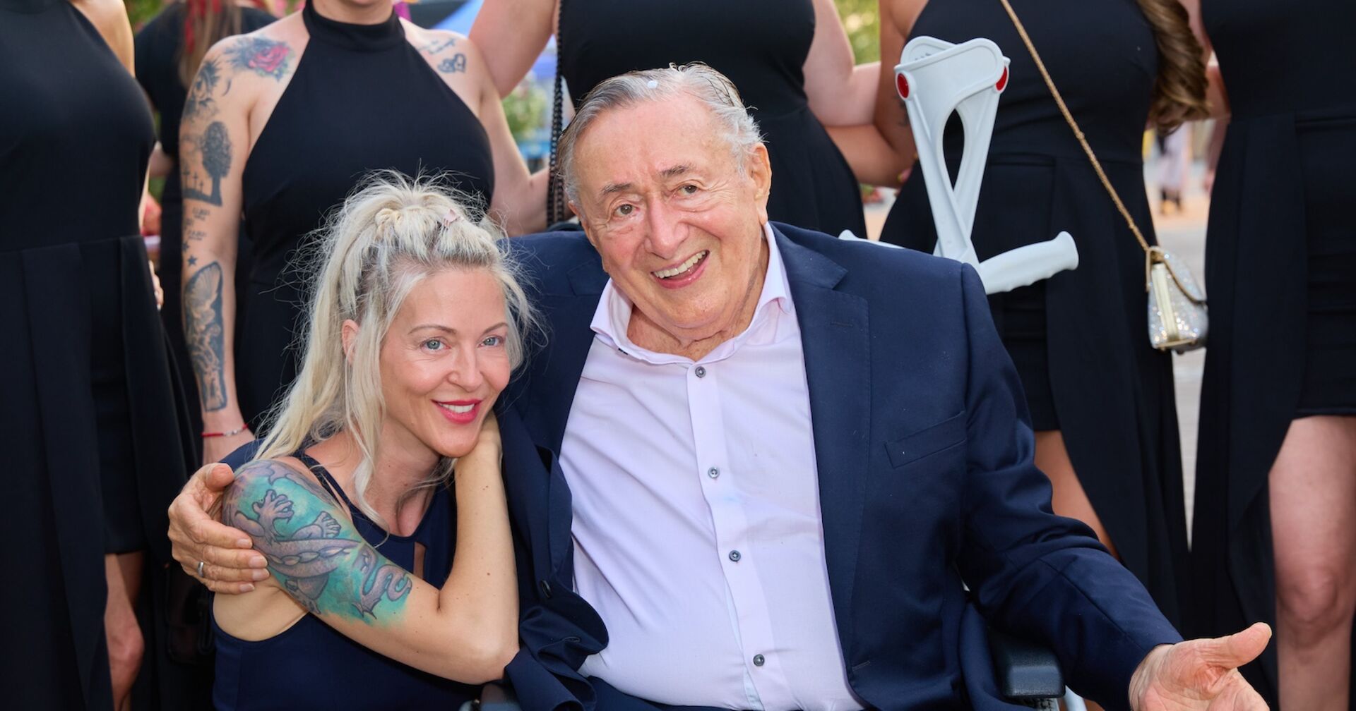 Simone kniet neben dem im Rollstuhl sitzenden Richard Lugner, er hat den Arm um sie gelegt. Beide lachen entspannt.