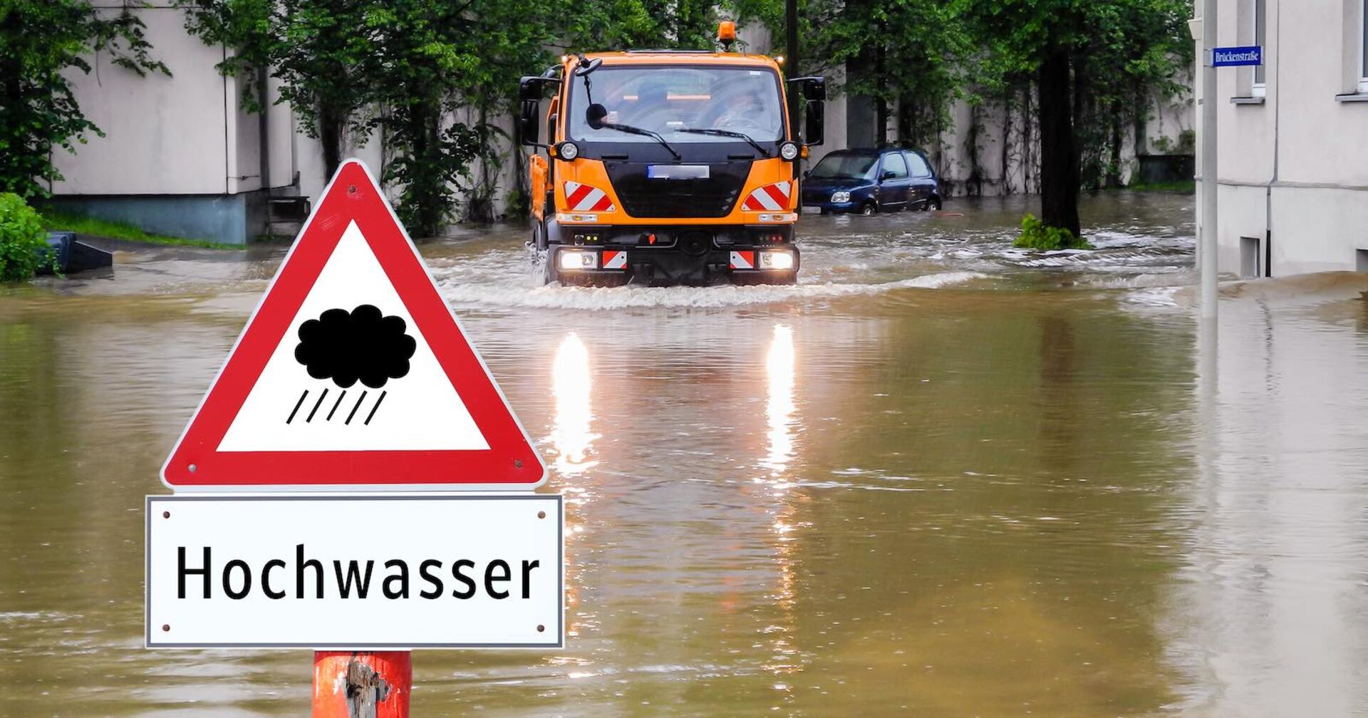 Hochwasser: Gemeinde-Lastwagen auf überschwemmter Straße und ein Hinweisschild &quot;Hochwasser&quot;.