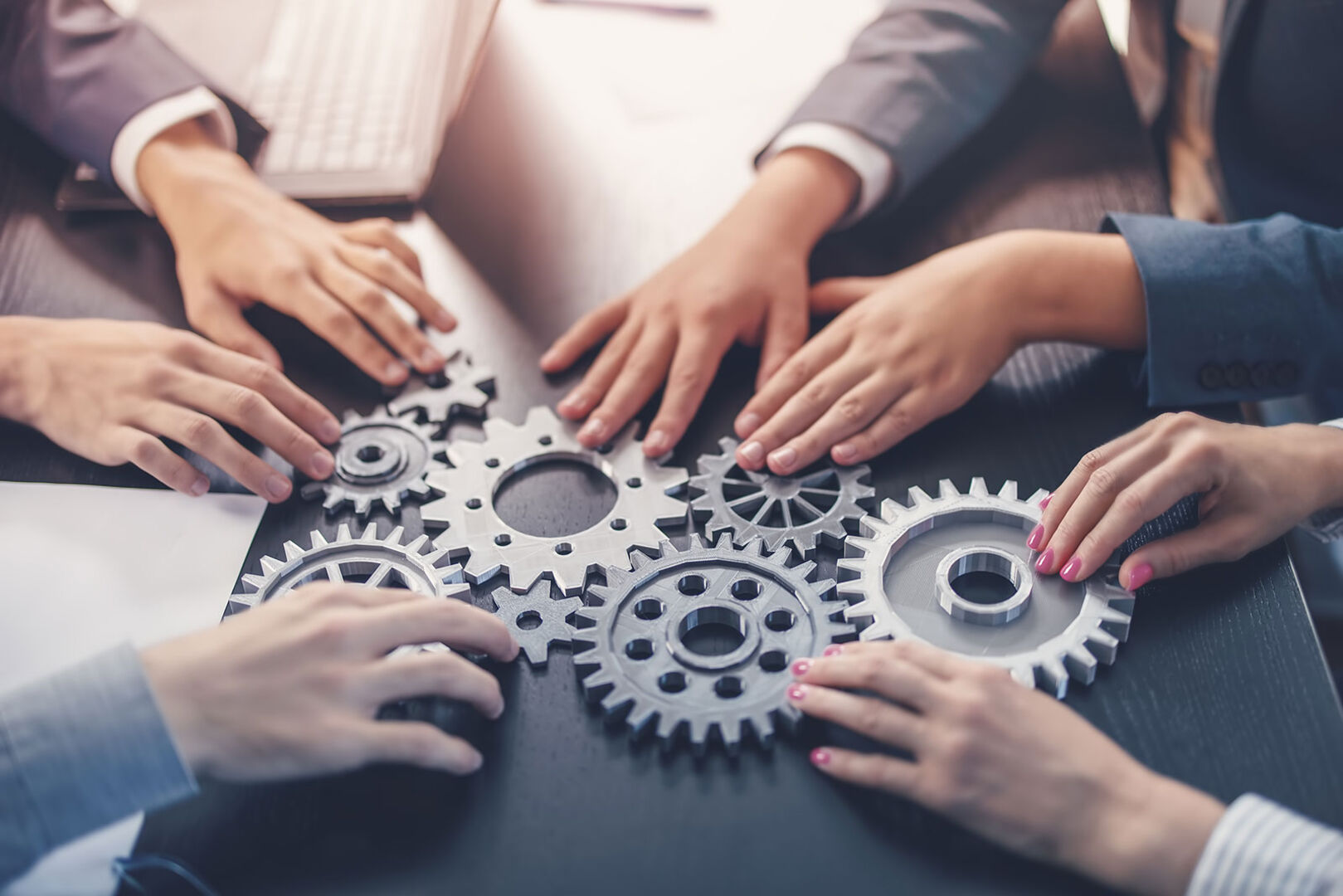 Frauen und Männer drehen am Bürotisch symbolisch an Zahnrädern | Credit: iStock.com/LeManna