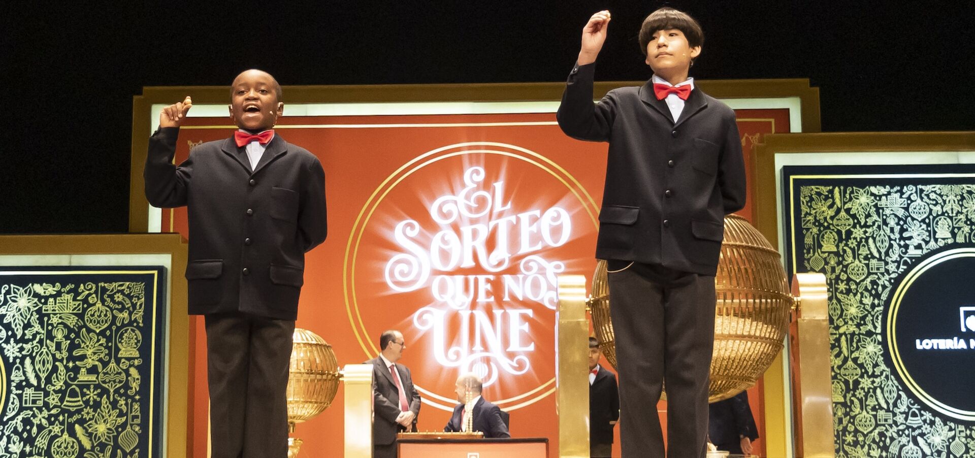 Zwei Burschen in Anzügen halten auf einer Bühne Losnummern hoch.