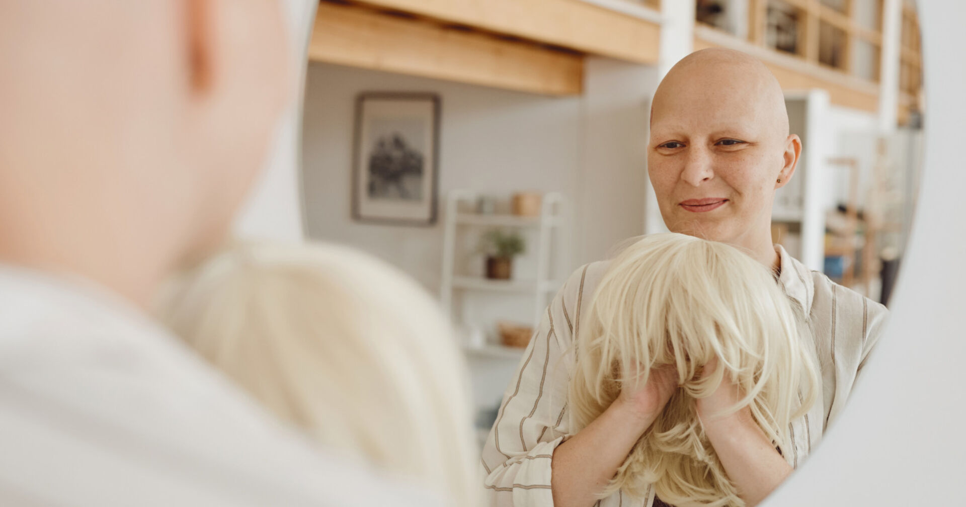 Frau mit Glatze hält Perücke in der Hand und schaut in einen Spiegel