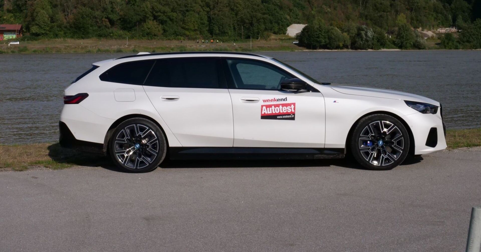 Ein weißer BMW i5 M60 xDrive  von der Seite. Das Auto leuchtet in der Sonne und steht am Ufer eines Flusses. Am anderen Ufer sind Wälder und Wiesen zu sehen.