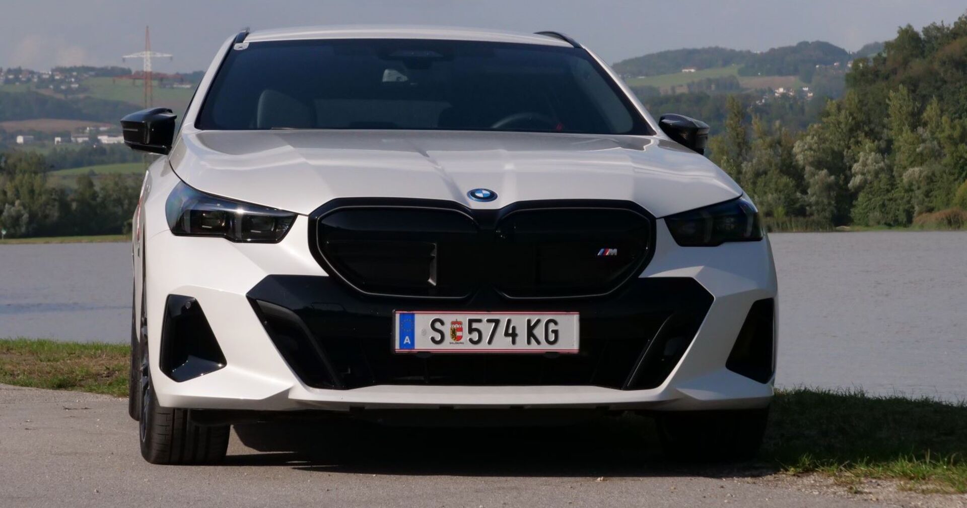 Ein weißer BMW i5 M60 xDrive - mit Salzburger Kennzeichen - steht am Ufer eines Flusses. Da Auto strahlt in der Sonne.